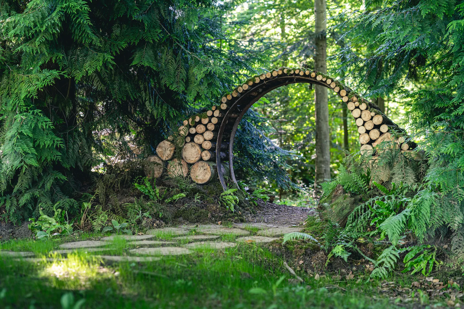 Une arche incurvée faite de rondins empilés se dresse dans une zone boisée au feuillage vert luxuriant et à la lumière du soleil.